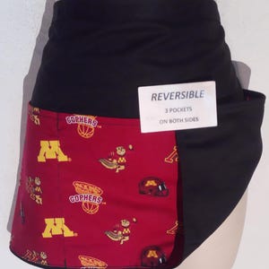 May include: A reversible apron featuring a black waistband and a maroon section printed with yellow Minnesota Gophers logos, basketball graphics, and helmets. A white tag attached to the front reads REVERSIBLE 3 POCKETS ON BOTH SIDES.