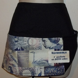 May include: Black reversible apron with a white and blue patterned fabric panel. The panel features a variety of images, including a woman's face, birds, and Japanese stamps. The apron has three pockets on both sides. The text "REVERSIBLE" and "3 pockets on BOTH sides" is printed on a white tag.