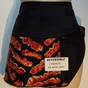 May include: Black reversible apron with a bacon print on one side and solid black on the other. The apron has three pockets on both sides. The text on the label reads "REVERSIBLE 3 POCKETS ON BOTH SIDES".