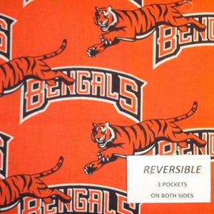 May include: Orange fabric with a repeating pattern of black and white Bengal tiger images and the word "Bengals" in a black and white arc. The fabric is reversible and has 3 pockets on both sides.