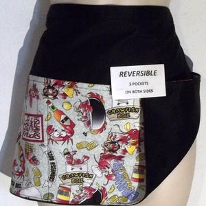 May include: A black and white reversible apron with a crawfish boil design. The apron has three pockets on both sides. The text "REVERSIBLE" and "3 POCKETS ON BOTH SIDES" is printed on a white tag attached to the apron.