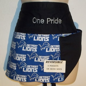 May include: A reversible black and blue apron with the Detroit Lions logo repeated on the blue side. The black side has the words "One Pride" embroidered in white. The apron has three pockets on each side and a tag that says "REVERSIBLE 3 POCKETS ON BOTH SIDES".