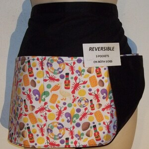 May include: A black and white reversible apron with a Mardi Gras theme. The apron has three pockets on each side and features a white background with a colorful pattern of beads, doubloons, hot sauce bottles, and fleur-de-lis. The text "REVERSIBLE" and "3 POCKETS ON BOTH SIDES" is printed on a white tag attached to the apron.
