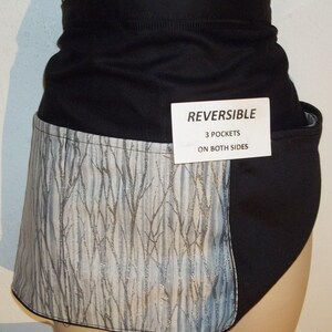 May include: Black and white reversible apron with a silver glitter tree pattern on one side and solid black on the other. The apron has three pockets on both sides. The text "REVERSIBLE 3 POCKETS ON BOTH SIDES" is printed on a white tag.