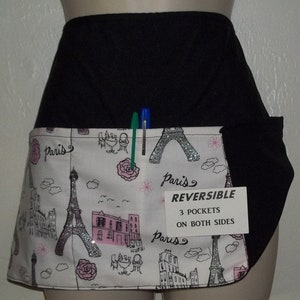 May include: A black and white reversible apron with a Paris theme. The apron has three pockets on both sides and features a silver glitter Eiffel Tower design. The text "Paris" and "REVERSIBLE" is printed on the apron.