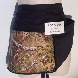 May include: Reversible black and camouflage apron with three pockets on both sides. The apron features a black waistband and a camouflage pattern with brown, green, and white accents. The tag reads "REVERSIBLE 3 POCKETS ON BOTH SIDES."