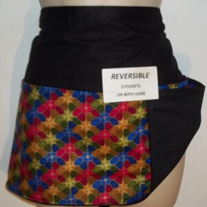 May include: A black and multi-colored reversible apron with three pockets on both sides. The apron has a geometric pattern with red, blue, green, and gold stars. The text "REVERSIBLE 3 POCKETS ON BOTH SIDES" is printed on a white tag.