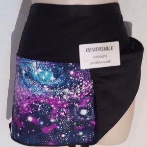 May include: Reversible black apron with a galaxy print on one side. The galaxy print features shades of blue, purple, and white. The apron has a tag that reads "REVERSIBLE 3 POCKETS ON BOTH SIDES."