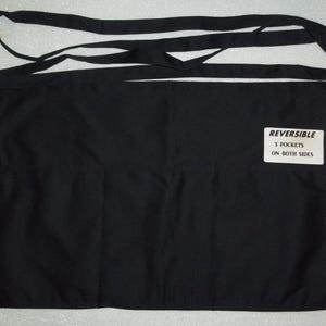 May include: Black reversible apron with three pockets on each side. The apron has a label that reads "REVERSIBLE 3 POCKETS ON BOTH SIDES".