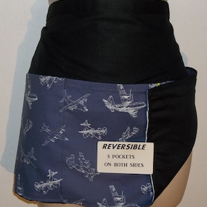 May include: A black and blue reversible apron with a white airplane pattern. The apron has three pockets on both sides. The text "REVERSIBLE 3 POCKETS ON BOTH SIDES" is printed on a white tag.