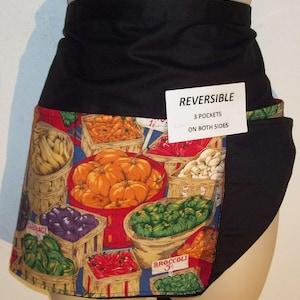 May include: Black reversible apron with a colorful fruit and vegetable print. The apron has three pockets on both sides. The text "REVERSIBLE" and "3 POCKETS ON BOTH SIDES" is printed on a white tag.
