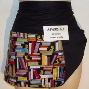 May include: Black reversible apron with a book print design. The apron has three pockets on both sides. The text "REVERSIBLE 3 POCKETS ON BOTH SIDES" is printed on a white tag.