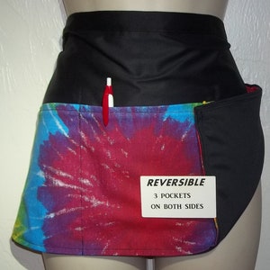 May include: Reversible black and tie-dye waist apron with three pockets on both sides. The tie-dye pattern features a vibrant mix of red, blue, purple, and yellow. A red and white pen is in the pocket.