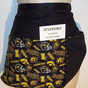 May include: Black and yellow reversible apron with three pockets on both sides. The apron features a repeating pattern of the University of Iowa Hawkeyes logo.