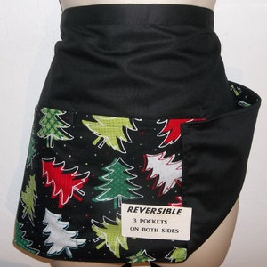 May include: Black reversible apron with a Christmas tree print on one side. The apron has three pockets on both sides. The text "REVERSIBLE 3 POCKETS ON BOTH SIDES" is printed on a white tag.