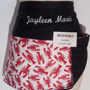 May include: Reversible black apron with a white and red lobster print on one side. The other side is black. The apron has the name "Jayleen Marie" embroidered in white script. A tag indicates the apron has three pockets on both sides.