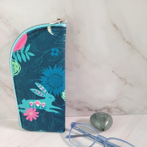 May include: A teal glasses case with a floral and bunny print, a blue pair of glasses, and a gray stone with the word "Love" engraved on it. The glasses case has a zipper closure and a striped trim.