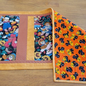 May include: A quilted Halloween table runner with a patchwork design. The center features panels with doll and pumpkin patterns. The border is orange, and the end has a repeating pattern of black cats and jack-o'-lanterns.