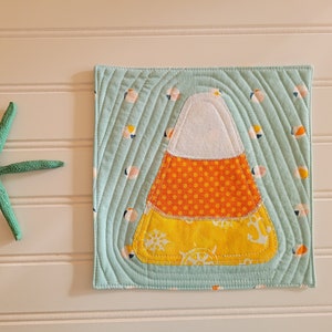 May include: A square, quilted fabric coaster featuring a candy corn design. The candy corn has white, orange polka dots, and yellow sections. The background is light blue with a nautical pattern. A green starfish is next to the coaster.