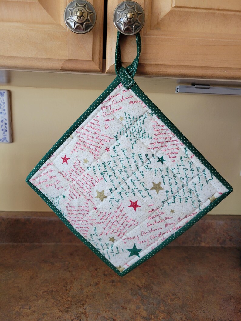 May include: A quilted Christmas pot holder with a green polka dot border and hanging loop. The white fabric features red and gold stars and the words "Merry Christmas".