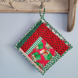 May include: A square, quilted Christmas potholder with a loop for hanging. The potholder features red fabric with small green Christmas trees, green and white striped fabric, and a central panel with green stockings on a red background.