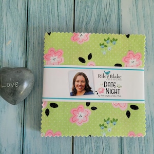May include: A square stack of light green fabric squares with a pink floral and black leaf pattern, wrapped in a white paper band labeled Riley Blake Designs Date Night by Keera Job of Fabric Mutt, next to a grey heart-shaped stone with the word Love.