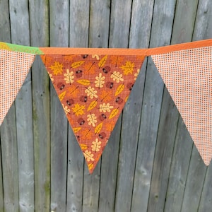 May include: A decorative pennant banner in autumnal colours. The central flag has a pattern of leaves and acorns on a brown background. The other flags have an orange polka dot pattern. The banner is strung on an orange ribbon.