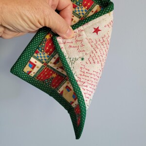May include: A Christmas-themed quilted pot holder with a green polka dot border. The interior has a festive pattern with red, gold, and blue elements, plus handwritten text. A green loop is attached for hanging.