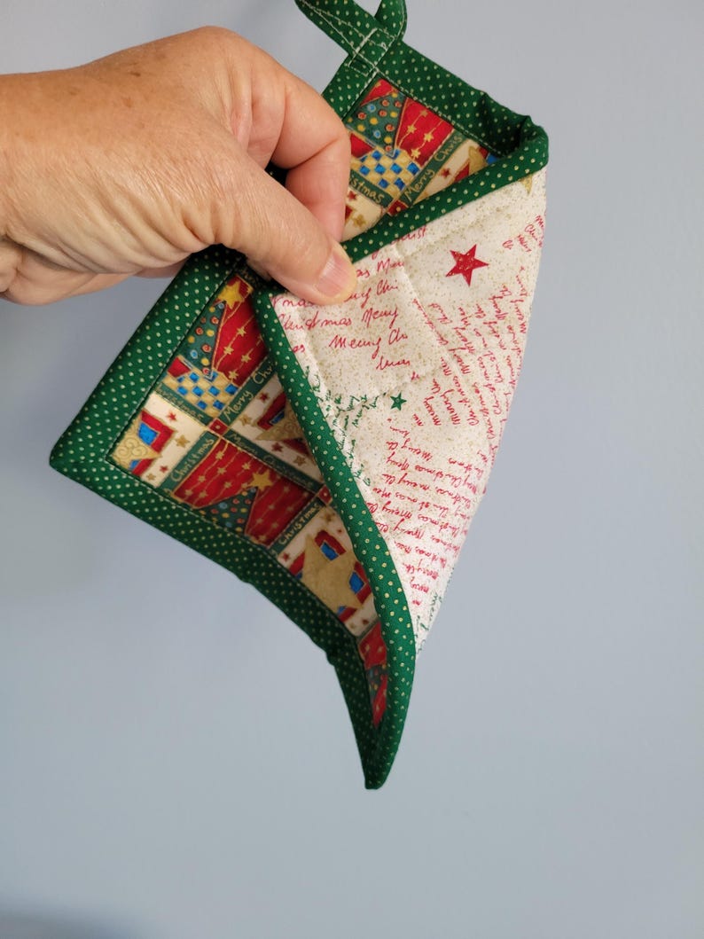 May include: A Christmas-themed quilted pot holder with a green polka dot border. The interior has a festive pattern with red, gold, and blue elements, plus handwritten text. A green loop is attached for hanging.