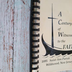 May include: A spiral-bound book with a black spine and cream-colored pages. The cover features a black illustration of a sailboat and the text "A Century of Witness to the Faith." The text "1895 Saint Ann Parish Wildwood, New Jersey" is also visible.