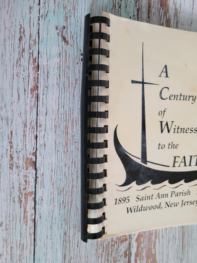May include: A spiral-bound book with a black spine and cream-colored pages. The cover features a black illustration of a sailboat and the text "A Century of Witness to the Faith." The text "1895 Saint Ann Parish Wildwood, New Jersey" is also visible.