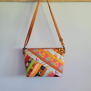 May include: A crossbody bag featuring a diagonal patchwork design with various floral, leaf, and animal patterns in shades of orange, yellow, blue, and green. The bag has a long, adjustable tan strap and a zipper closure along the top edge.