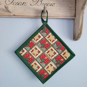 May include: A quilted, square hot pad with a green border and hanging loop. The center features a repeating pattern of red, gold, and blue Christmas-themed designs, including stars and trees. The hot pad is hanging on a wooden rack.