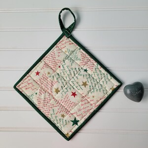 May include: A square Christmas pot holder with a green border and loop. The cream fabric is patterned with red and green script and gold and red stars. The pot holder is quilted and has a green hanging loop.