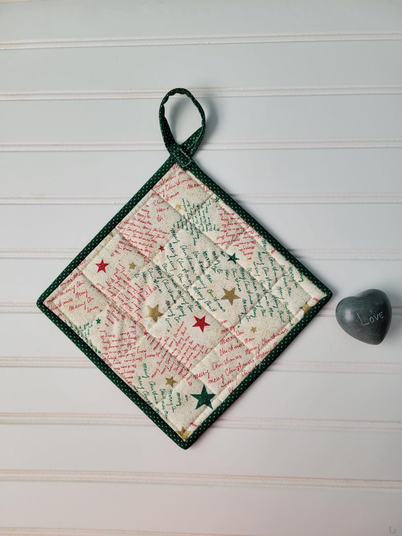 May include: A square Christmas pot holder with a green border and loop. The cream fabric is patterned with red and green script and gold and red stars. The pot holder is quilted and has a green hanging loop.