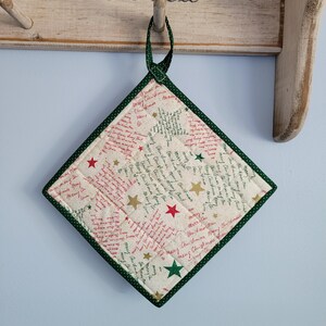 May include: A square Christmas-themed quilted potholder with a green border and hanging loop. The off-white fabric is patterned with red and green text, gold and red stars. The potholder is hanging on a wooden rack.