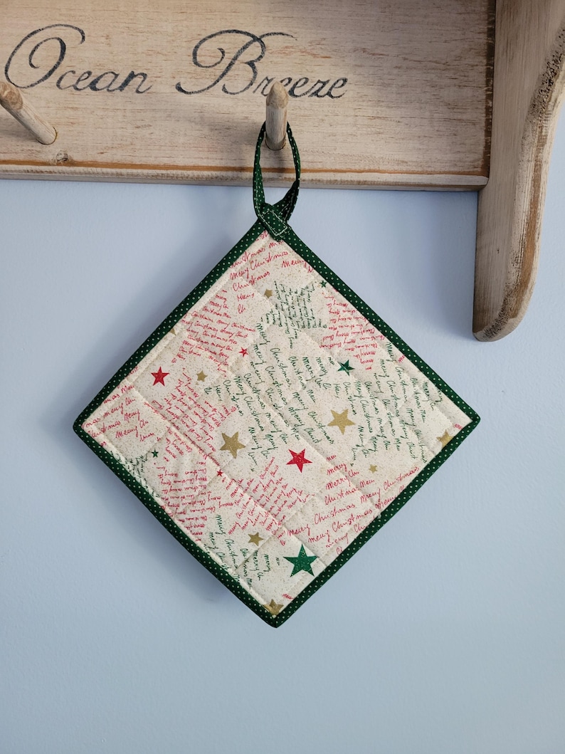 May include: A square Christmas-themed quilted potholder with a green border and hanging loop. The off-white fabric is patterned with red and green text, gold and red stars. The potholder is hanging on a wooden rack.