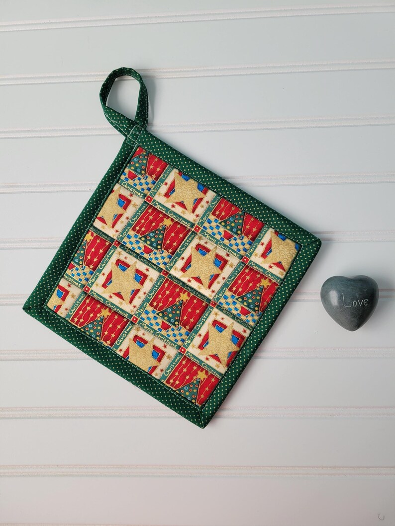 May include: Square Christmas potholder with a green border and hanging loop. The fabric has a repeating pattern of Christmas trees, stars, and the words "Merry Christmas" in red, blue, and gold. A small grey heart-shaped stone is next to the potholder.
