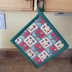 May include: A festive square potholder with a green polka dot trim and loop. The central design is a patchwork of red, blue, and gold star and text patterns. The text reads "Merry Christmas".