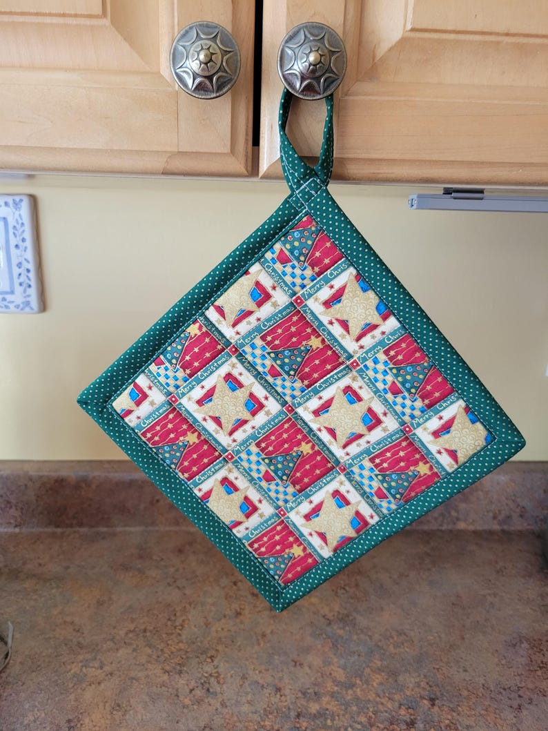 May include: A festive square potholder with a green polka dot trim and loop. The central design is a patchwork of red, blue, and gold star and text patterns. The text reads "Merry Christmas".