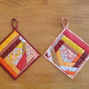 May include: Two handmade, square quilted potholders. One features a fox design and warm colors like orange, yellow, and burgundy. The other has a similar design with a different fox and a lighter color palette. Both have hanging loops.