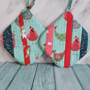 May include: Two quilted pot holders with a whimsical chicken design. The pot holders feature a light teal background with vertical stripes of red, pink, and floral patterns. Each has a loop for hanging.