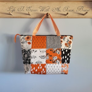 May include: A patchwork tote bag with a variety of patterns, including orange polka dots, black and white floral, and a checkered design. The bag has orange and white striped handles and a zipper closure. The background includes a wooden shelf with the text "Life Is Easier With An Ocean Breeze."