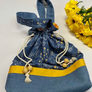 May include: A blue drawstring bag with a yellow ribbon and white floral print. The bag has a long blue strap and wooden beads on the drawstring.
