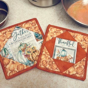 May include: Two quilted pot holders with an autumn theme. One pot holder has a brown background with a white pumpkin and the text "thankful". The other pot holder has a white background with a pumpkin and the text "gather here with grateful hearts".