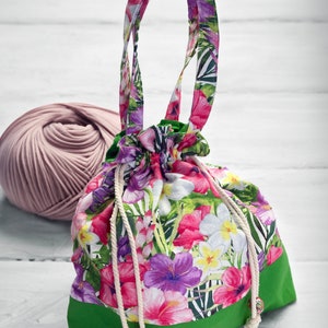 May include: A green and white drawstring bag with a floral print. The bag has a green bottom and a white drawstring with wooden beads. The bag is made of fabric and has a floral print with pink, white, and purple flowers.