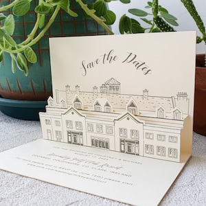 May include: A cream-colored pop-up card featuring a line-art illustration of a building. The text reads Save the Dates, For the wedding of Courtney & Michael, August 17, 2023, Doonbeg Golf Club Resort, Doonbeg, Doughmore, Co. Clare, Ireland.