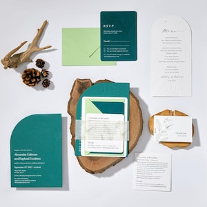 May include: An assortment of teal and green wedding stationery, including invitations, RSVP cards, and a menu. The invitations feature the names "Alexandra & Raphael" and the wedding date. The stationery is arranged with natural wood and pinecones.
