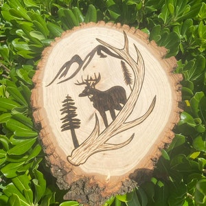 May include: A wood slice with a burned design of a deer with antlers, a mountain range, and a pine tree.
