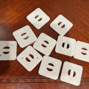 May include: A collection of ten white square plastic buttons, each featuring rounded corners and a central horizontal slot flanked by two small circular holes. The buttons are arranged in a scattered pattern, displaying a smooth, flat surface.